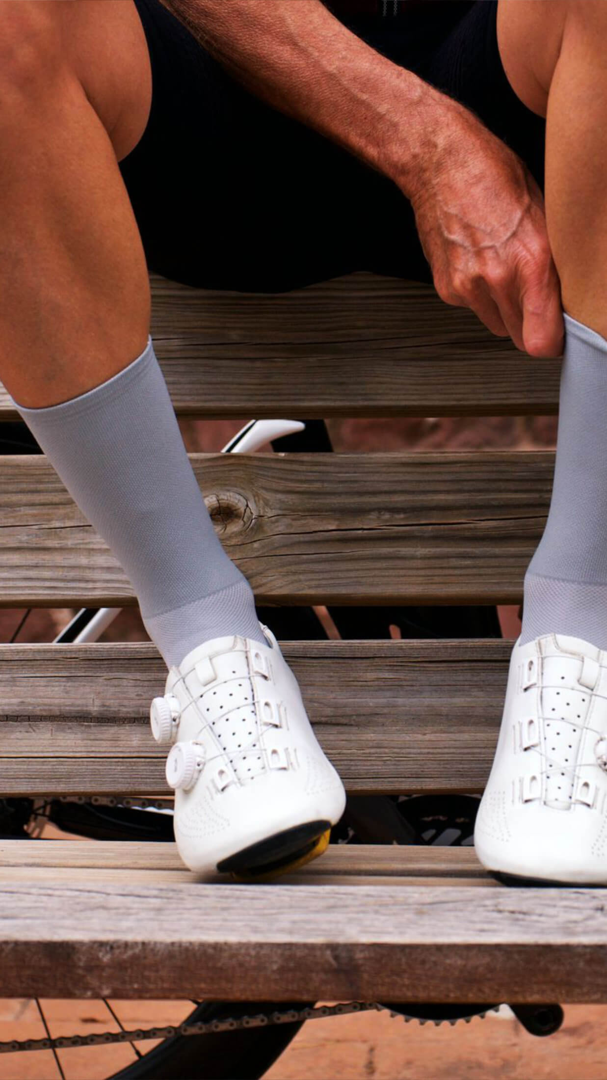 Close-up of a seated cyclist adjusting nologo classic gray cycling socks while wearing white cycling shoes, seated on a wooden bench.