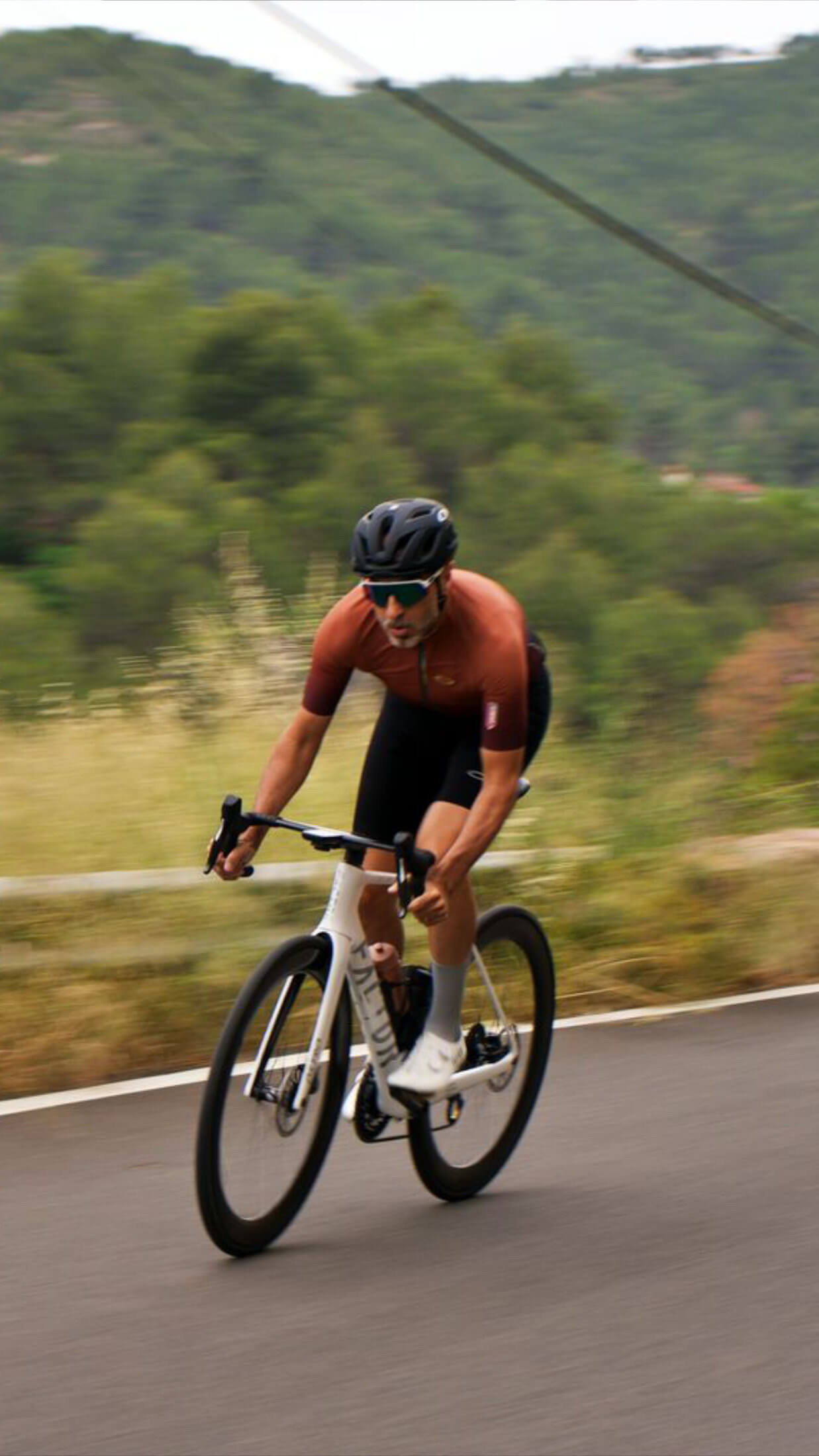 Cyclist wearing nologo classic gray cycling socks, brown jersey, and black bibs riding at speed on a curvy mountain road.