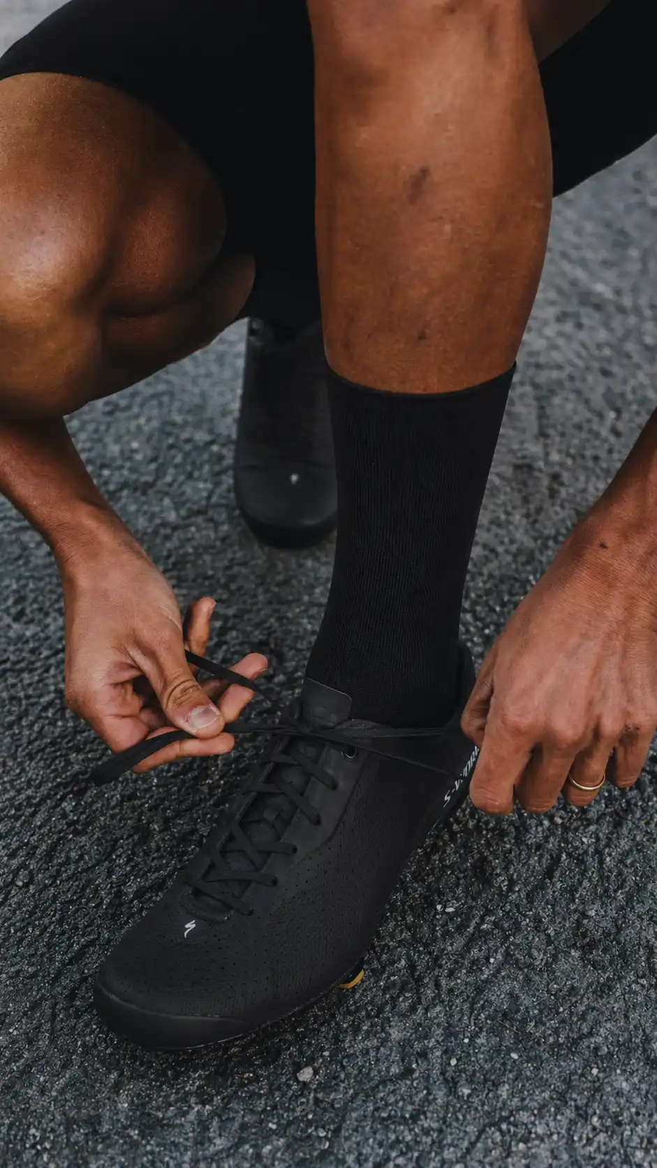Close-up of a cyclist tying black road cycling shoes while wearing black nologo cycling socks on wet asphalt.