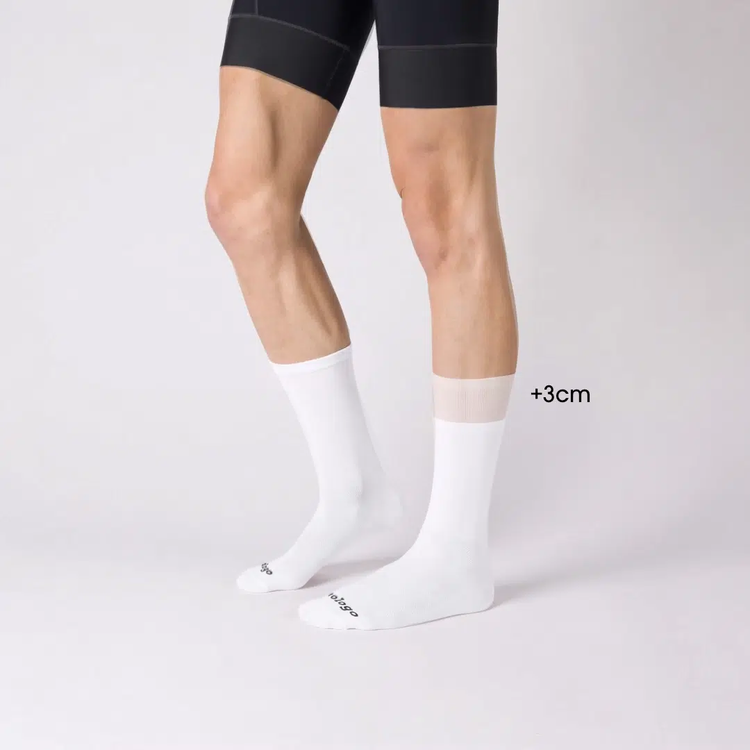 Vista laterale di calze da ciclismo lunghe bianche senza logo con un confronto visivo di +3 cm di altezza, che mostra la lunghezza del bordo esteso rispetto alla versione standard.